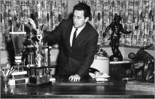 Joe Weider behind a desk surrounded by trophies