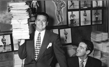 Two suited men in an office, one holding a tall stack of papers